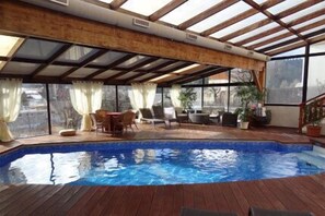 An indoor swimming pool with wooden decking, a covered seating area with chairs and a table, and large windows with curtains. - Le Christiania Hotel (Villard-de-Lans)