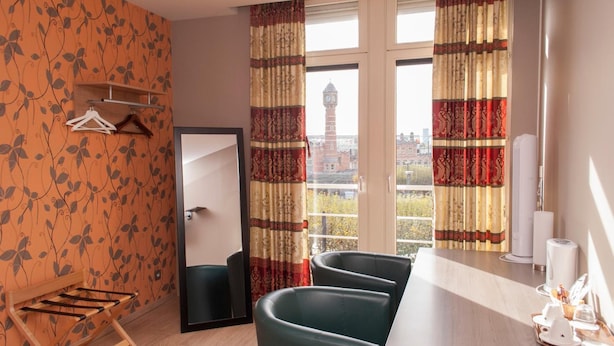A hotel room with a large window, a wooden drying rack, two green chairs, a small table with toiletries, and a view of a clock tower. - Hotel Castel (Ghent)