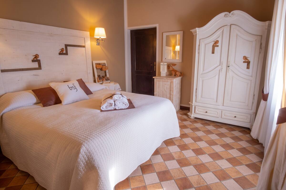 Een keurig opgemaakt bed met een witte deken en een kussen met 'b&f' geborduurd erop. Daarbij staat een klein tafeltje met een lamp en een ingelijste foto. Tegen de muur bevindt zich een witte garderobe met een spiegel, en er is een deur te zien op de achtergrond.