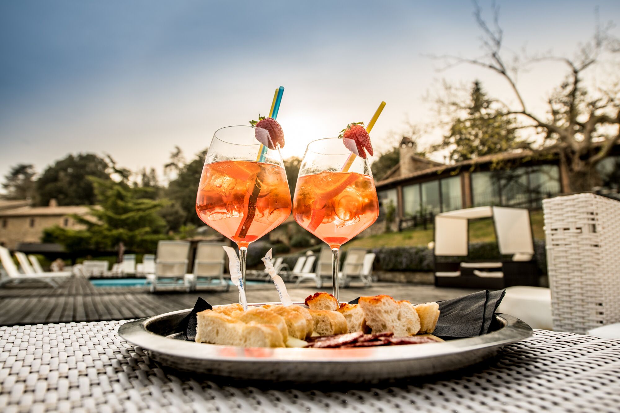 Dois copos de cocktail rosa com guarnições de fruta sobre uma mesa redonda com um prato de pão e queijo.