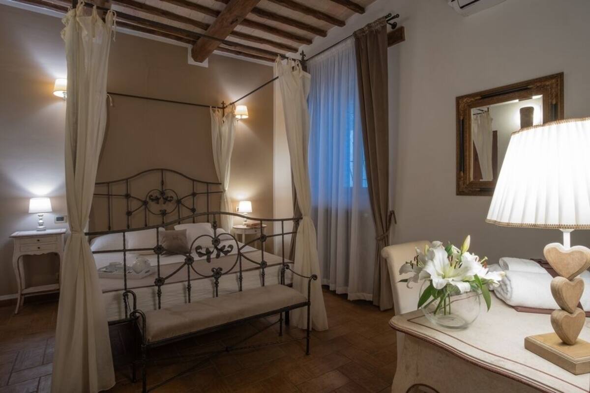 Een slaapkamer met een hemelbed, een nachtkastje met een lamp, een stoel en een nachtkastje met een bloemenvaas.