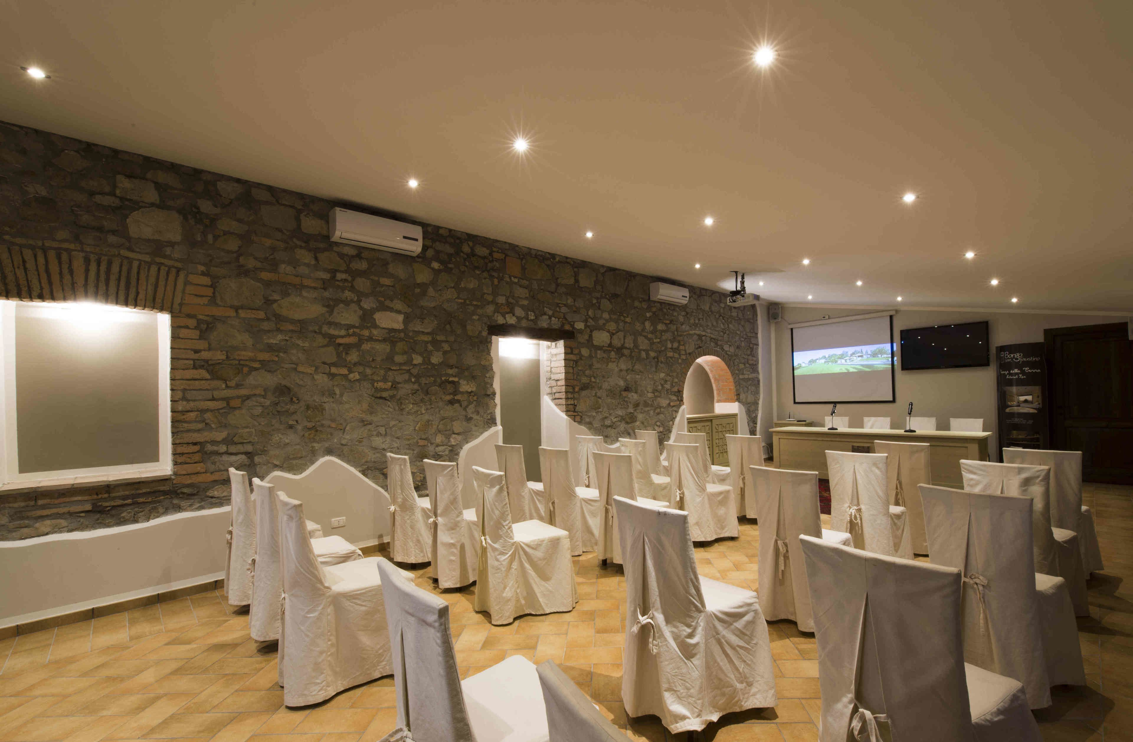 A conference room with stone walls, a projector screen, and rows of chairs covered with white cloths.