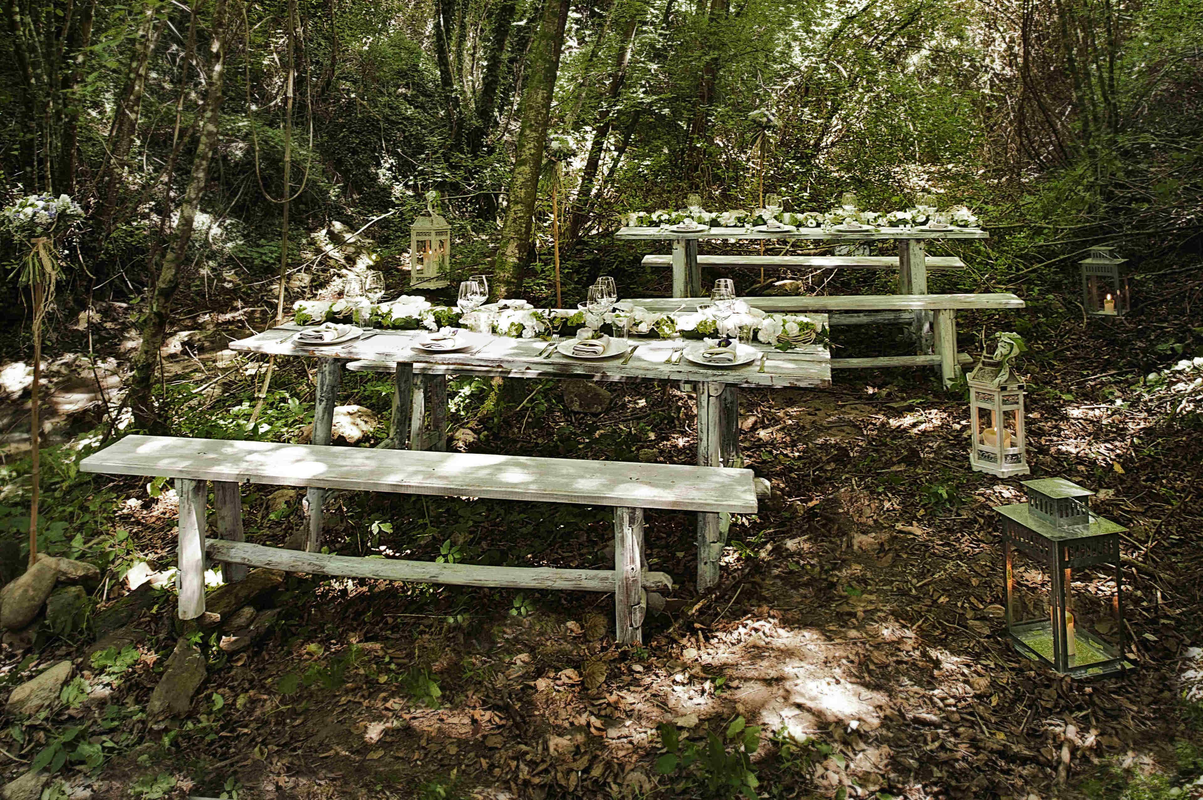 A rustic wooden picnic table set up in a forest with white flowers and lanterns.