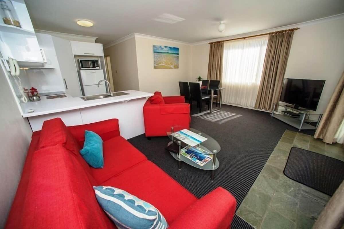 Ein modernes Wohnzimmer mit einem roten Sofa, einem Glastisch, einem Flachbildfernseher und einer Küchenzeile mit weißen Schränken und einer Mikrowelle.
