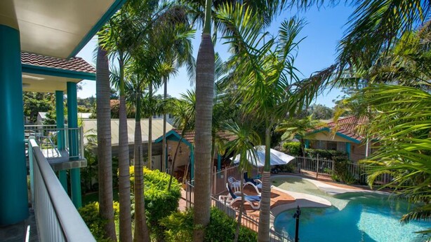 Un área de piscina con palmeras, sillas de descanso y una zona de estar cubierta.