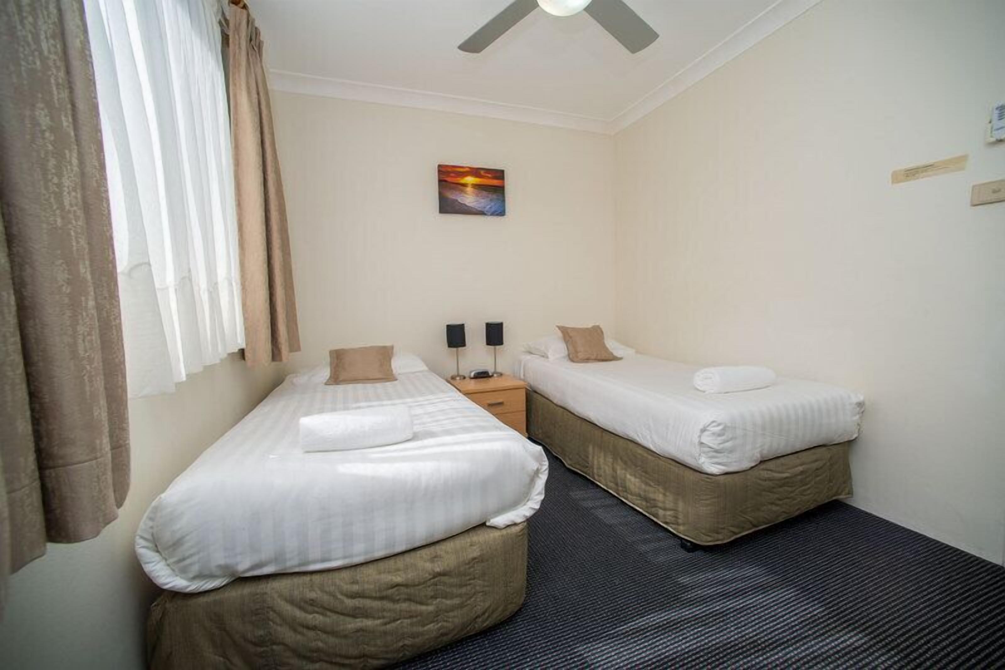 Een hotelkamer met twee eenpersoonsbedden, een plafondventilator, een schilderij aan de muur en een raam met gordijnen.