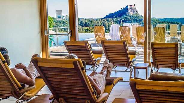 A sunlit room with wooden chairs and a view of a hill with a castle. - Genusshotel Riegersburg (Riegersburg)