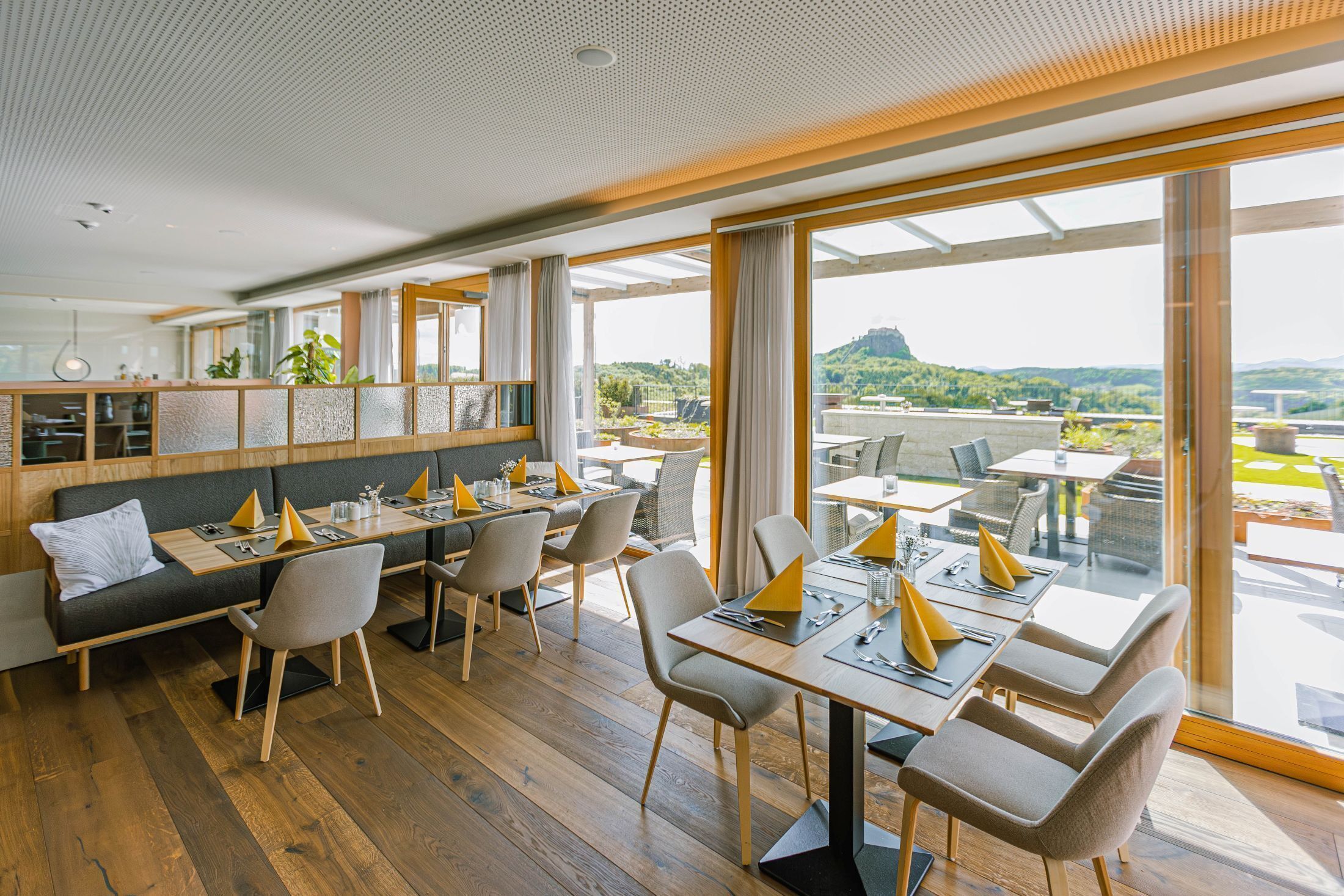 A modern dining area with wooden flooring, grey upholstered seating, and large windows offering a view of a mountainous landscape.