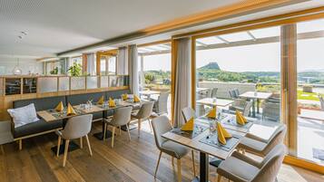 A modern dining area with wooden flooring, grey upholstered seating, and large windows offering a view of a mountainous landscape.