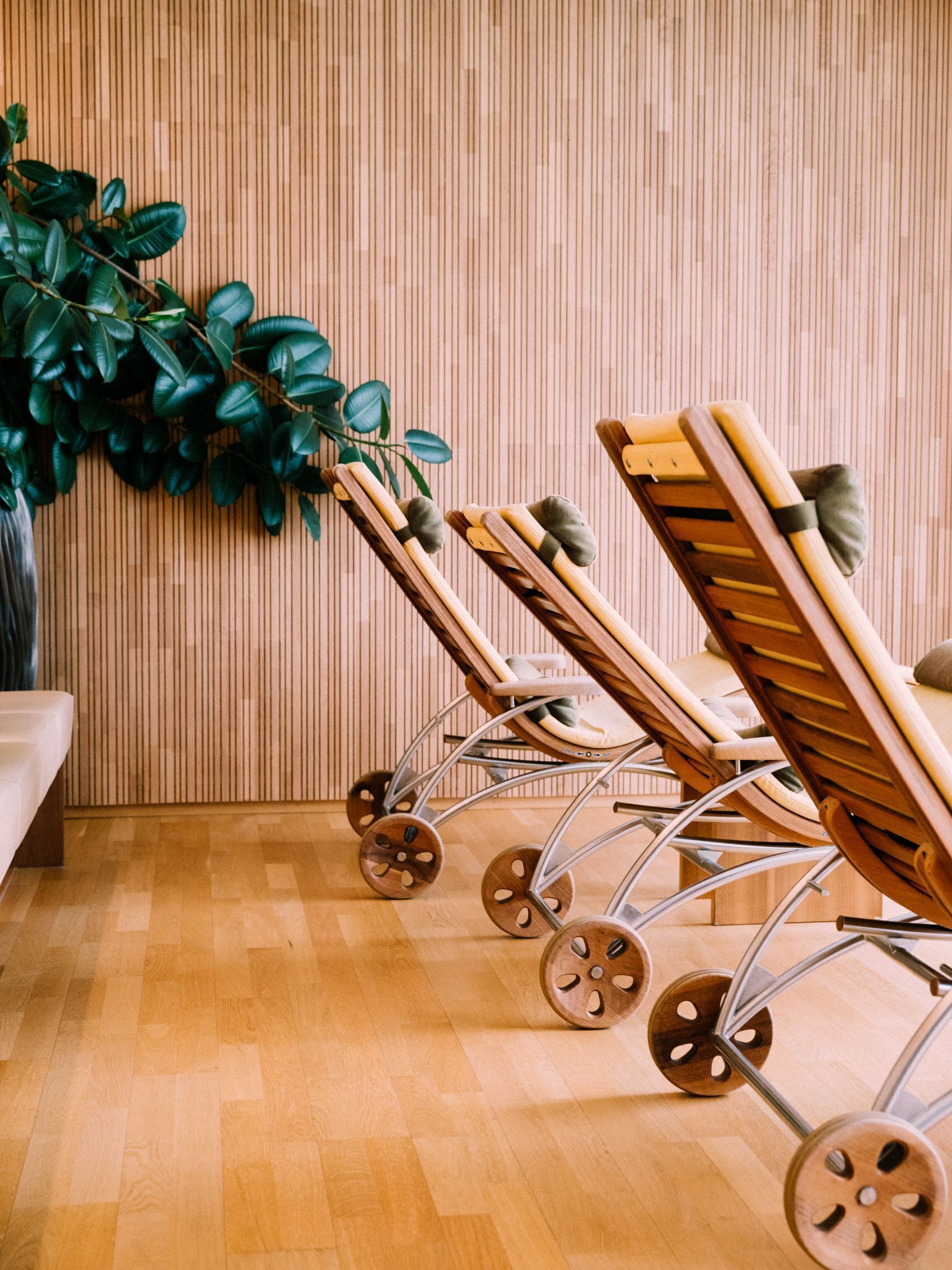 A row of wooden lounge chairs with metal wheels, positioned against a wooden wall with vertical lines.
