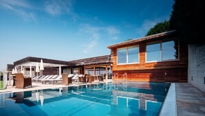 A swimming pool with lounge chairs and umbrellas, a wooden building with large windows, and a clear blue sky. - Genusshotel Riegersburg (Riegersburg)
