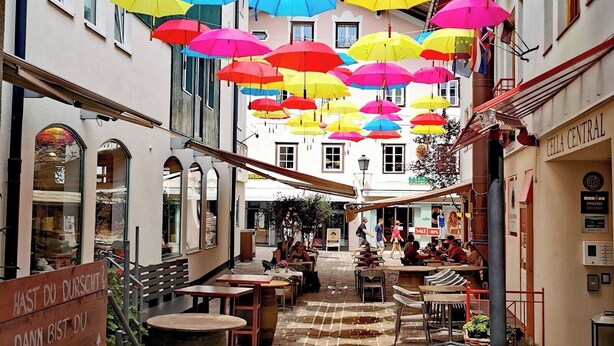 Ein Innenhof mit bunten Sonnenschirmen, Sitzgelegenheiten im Freien und einem Schild auf Deutsch.