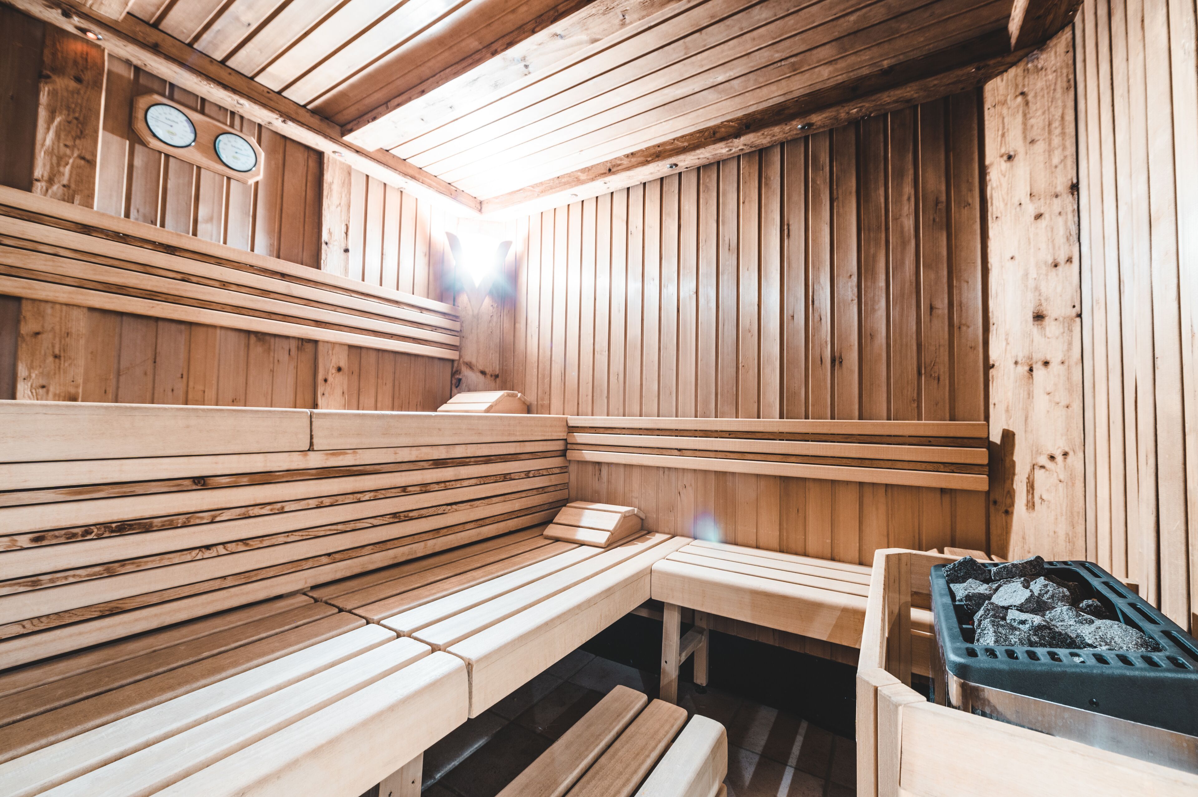 A sauna with wooden benches and a bench seat, a wooden wall with a clock, and a container with stones.