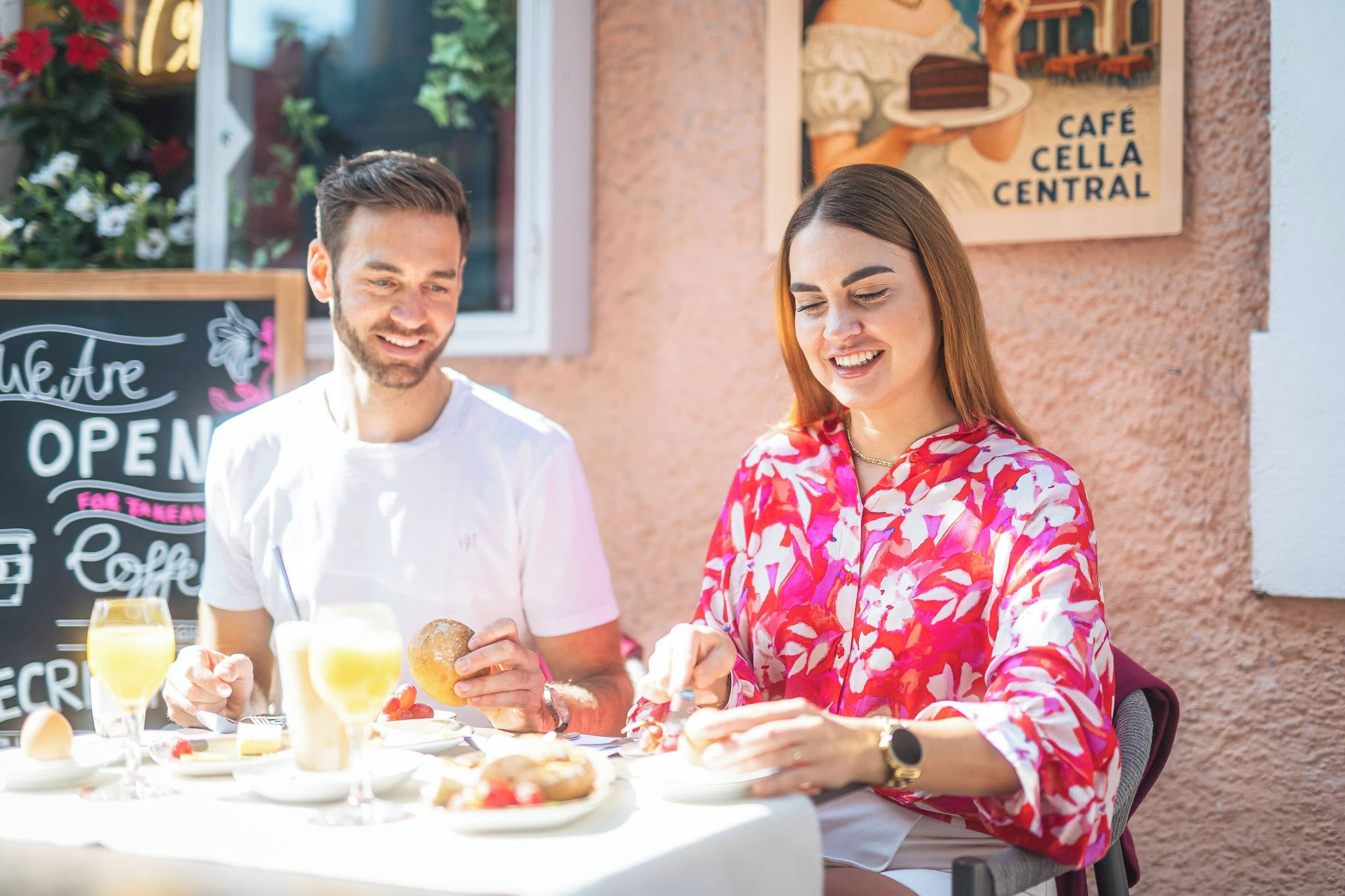 Una pareja cenando en un café al aire libre, con un cartel de pizarra que indica que el establecimiento está abierto para el desayuno.