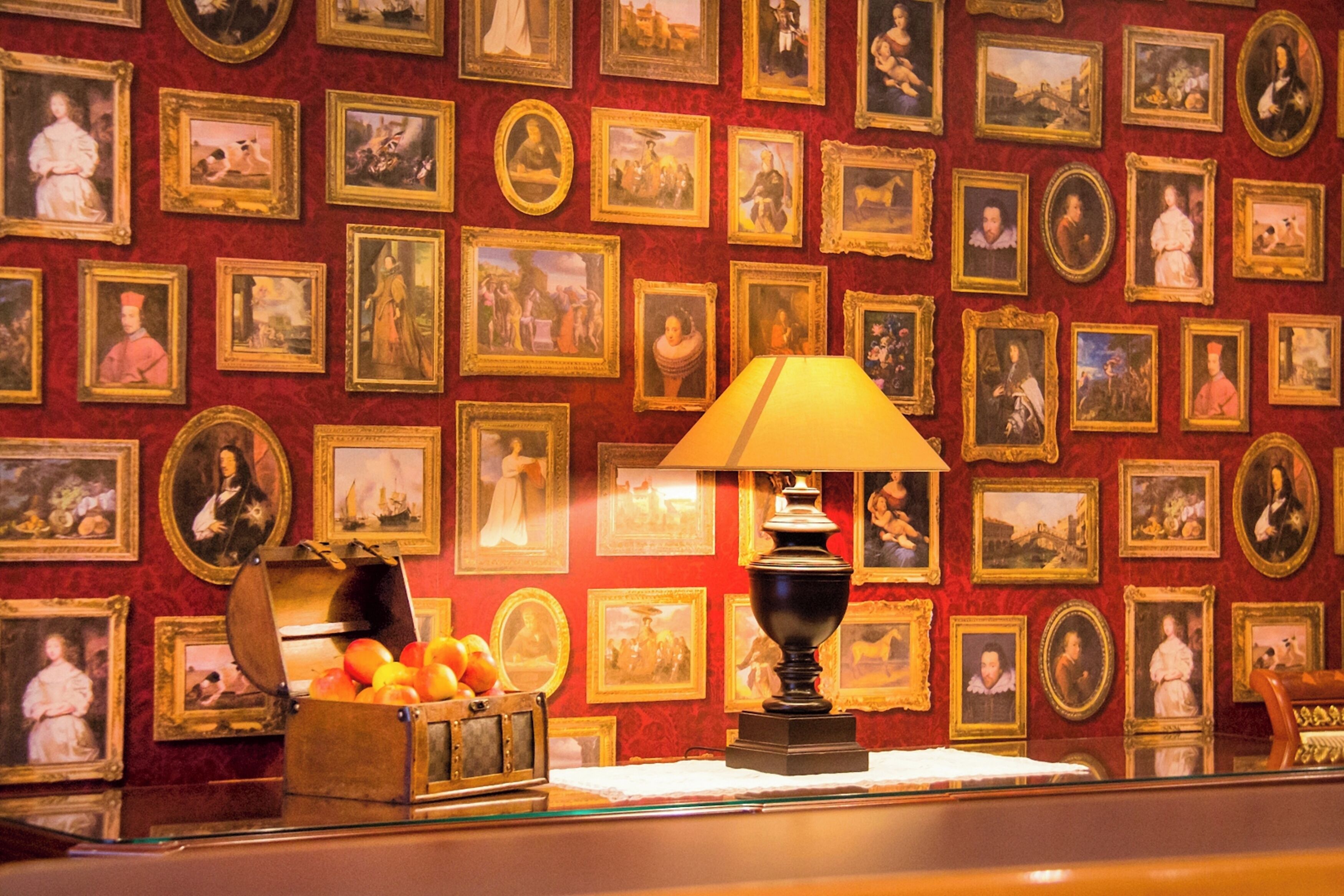 A wall with numerous framed portraits and a table with a lamp and fruit basket.