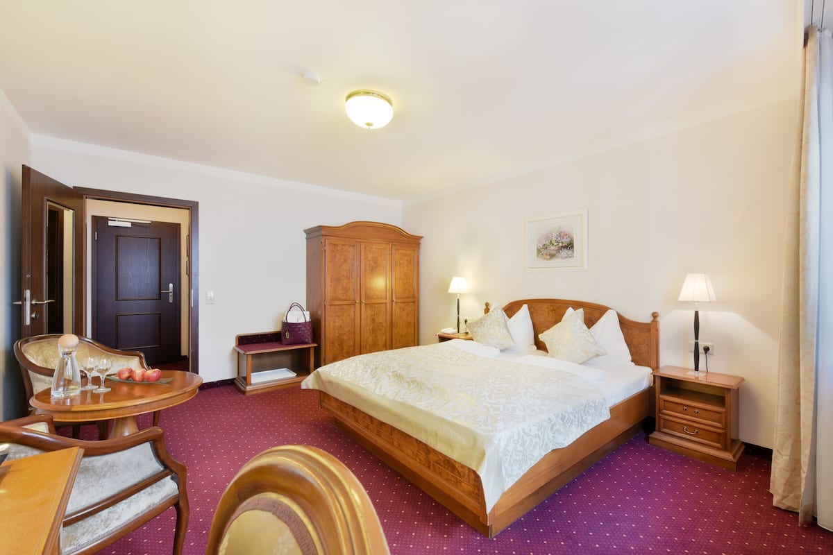 Une chambre d’hôtel avec un grand lit, une armoire en bois, une petite table avec une chaise et un bureau avec une lampe.
