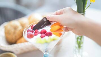 A hand holding a card with a heart symbol, a dessert with fruits, a basket of bread, and a vase with a yellow flower.