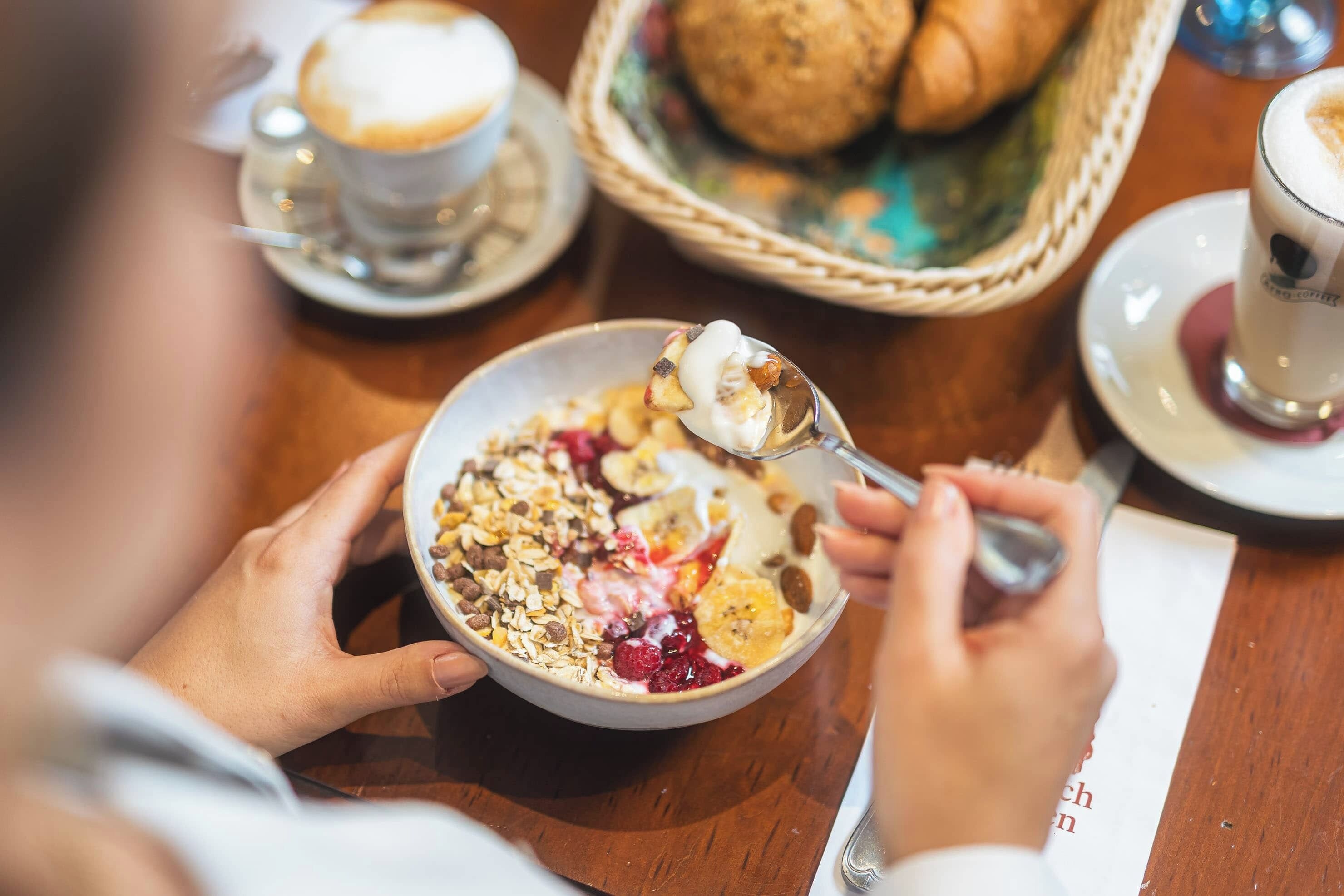 Eine Person isst eine Schüssel Müsli mit Früchten und Nüssen, eine Tasse Kaffee und einen Korb Brot an einem Holztisch.