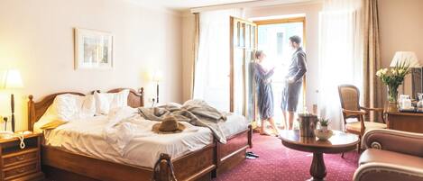 A hotel room with a large bed, a wooden nightstand, a desk with a lamp, a chair, and a small table with a vase of flowers.