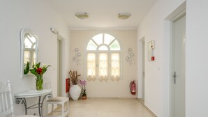 A hallway with a white door, a white bench, a table with a vase of flowers, and a large window with curtains. - Golden Beach Studios (Naxos)