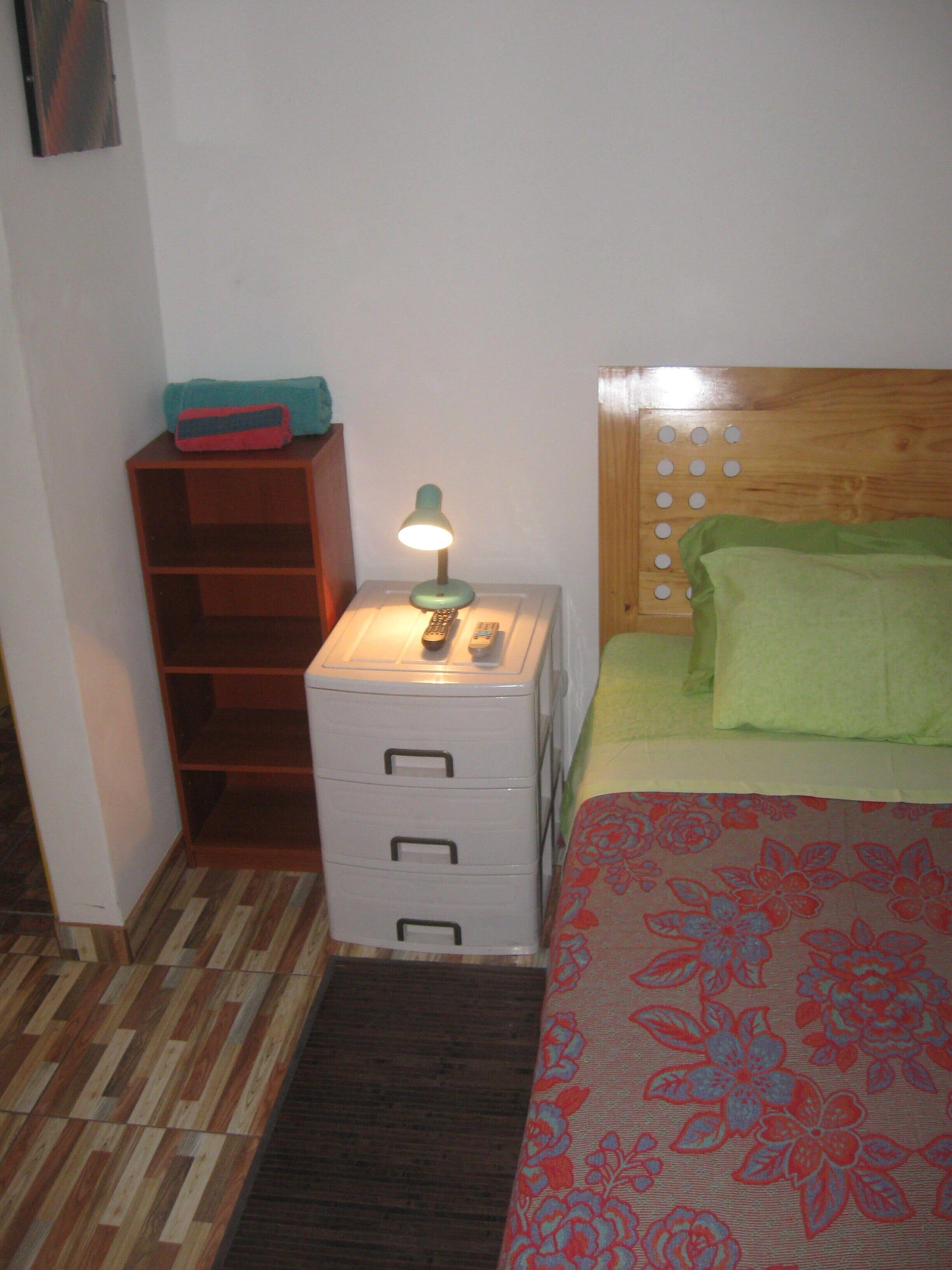Ein kleines, ordentlich eingerichtetes Schlafzimmer mit einem Holzkopfende, einer Nachttischlampe, einem Nachttisch mit Schubladen und einer gemusterten Bettdecke.