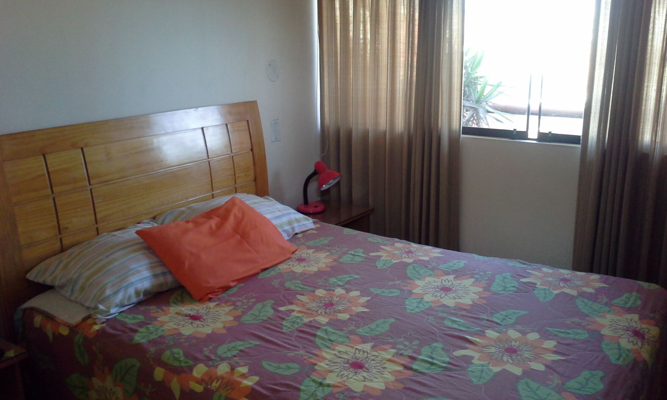 Una cama con cubrecama floral, cabecera de madera y una lámpara roja sobre una mesita de noche.