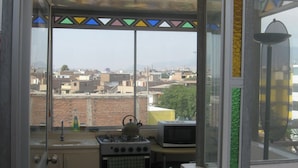 A small kitchen with a gas stove, a microwave, and a view of the city through a window with colorful stained glass. - Posada Peregrinus (Lima)