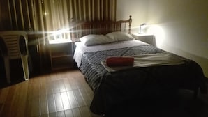 A small, dimly lit room with a single bed, a nightstand with a lamp, a chair, and a window with curtains. - Posada Peregrinus (Lima)