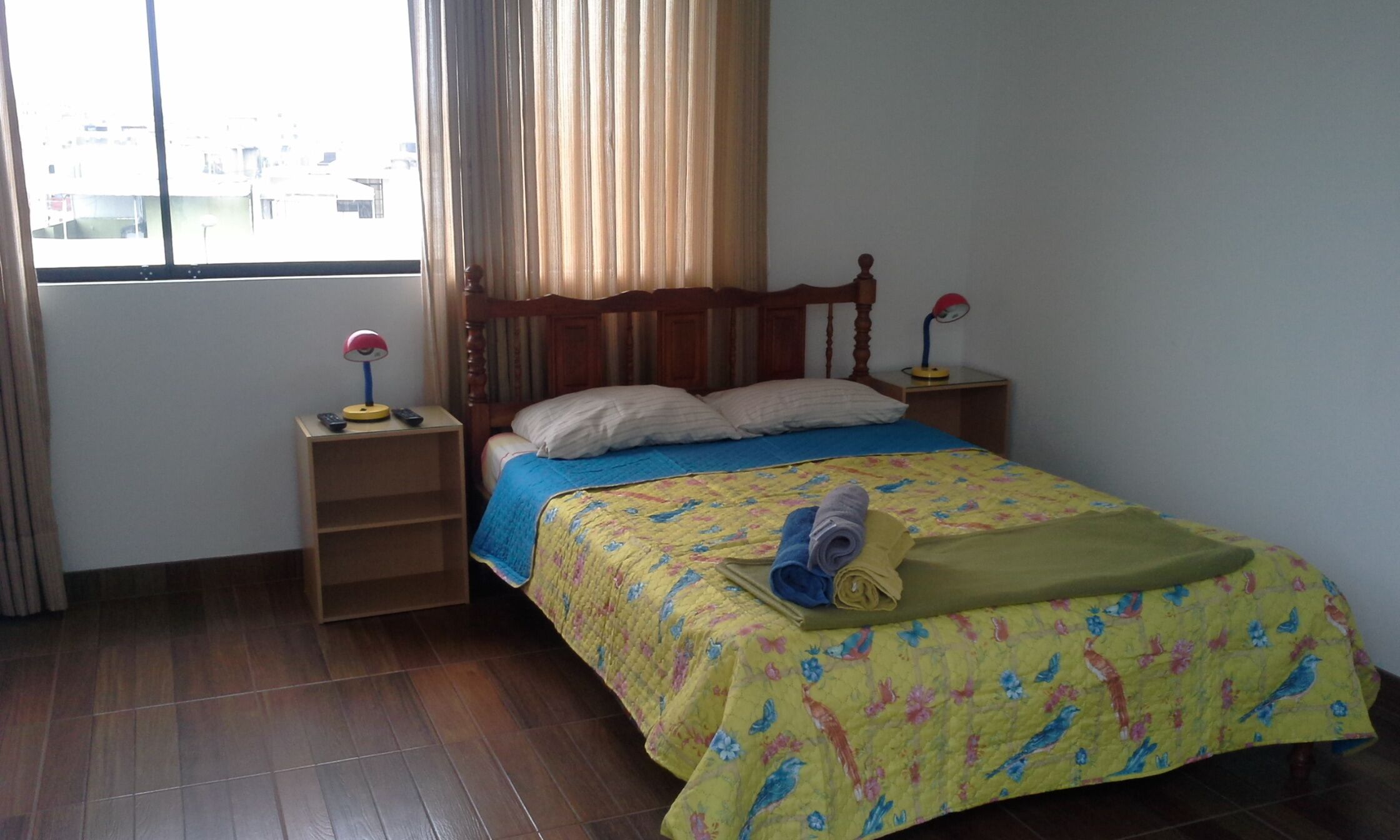 A bedroom with a wooden bed, two bedside lamps, a window with curtains, and a wooden floor.