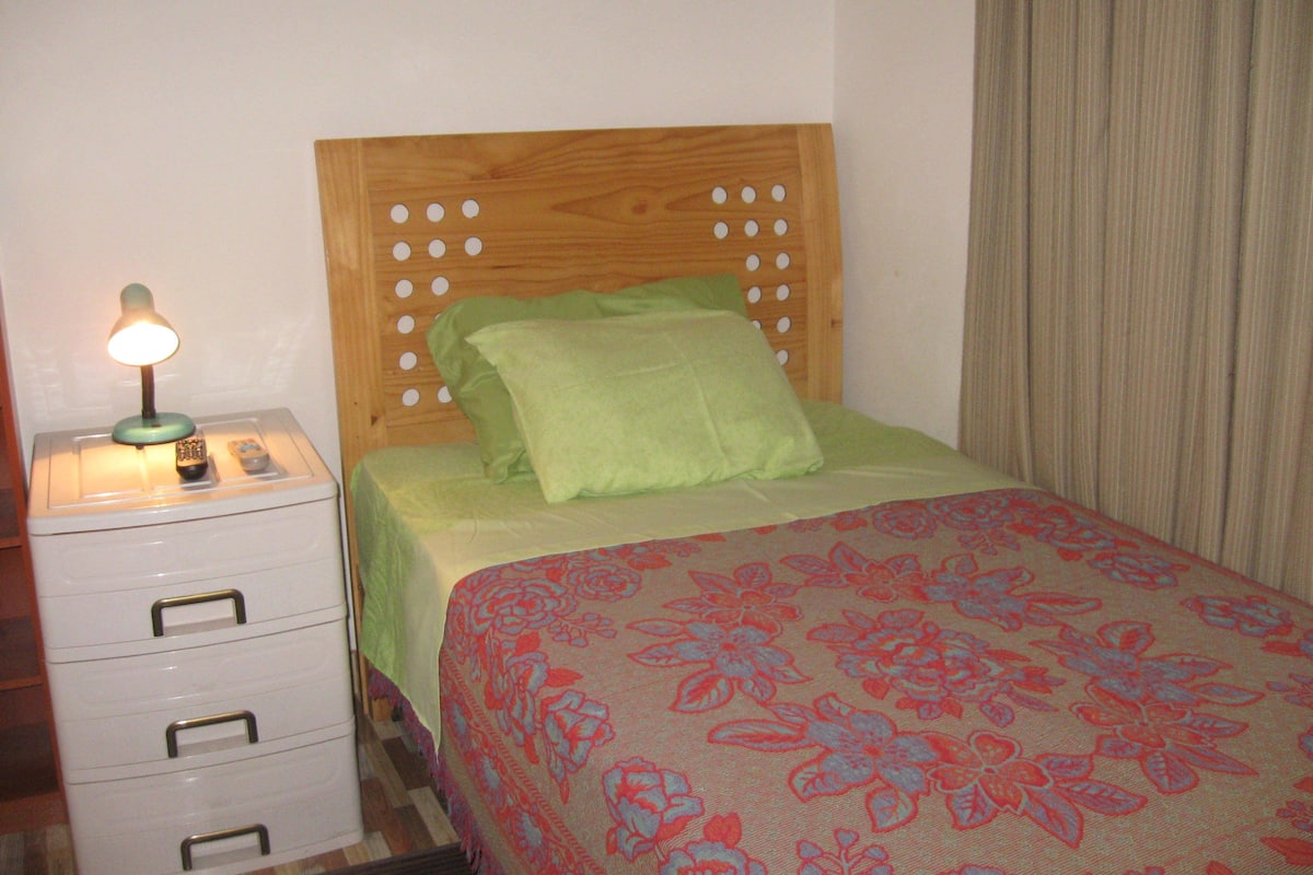 A bedroom with a wooden headboard, a green pillow, a floral bedspread, and a bedside table with a lamp and remote controls.