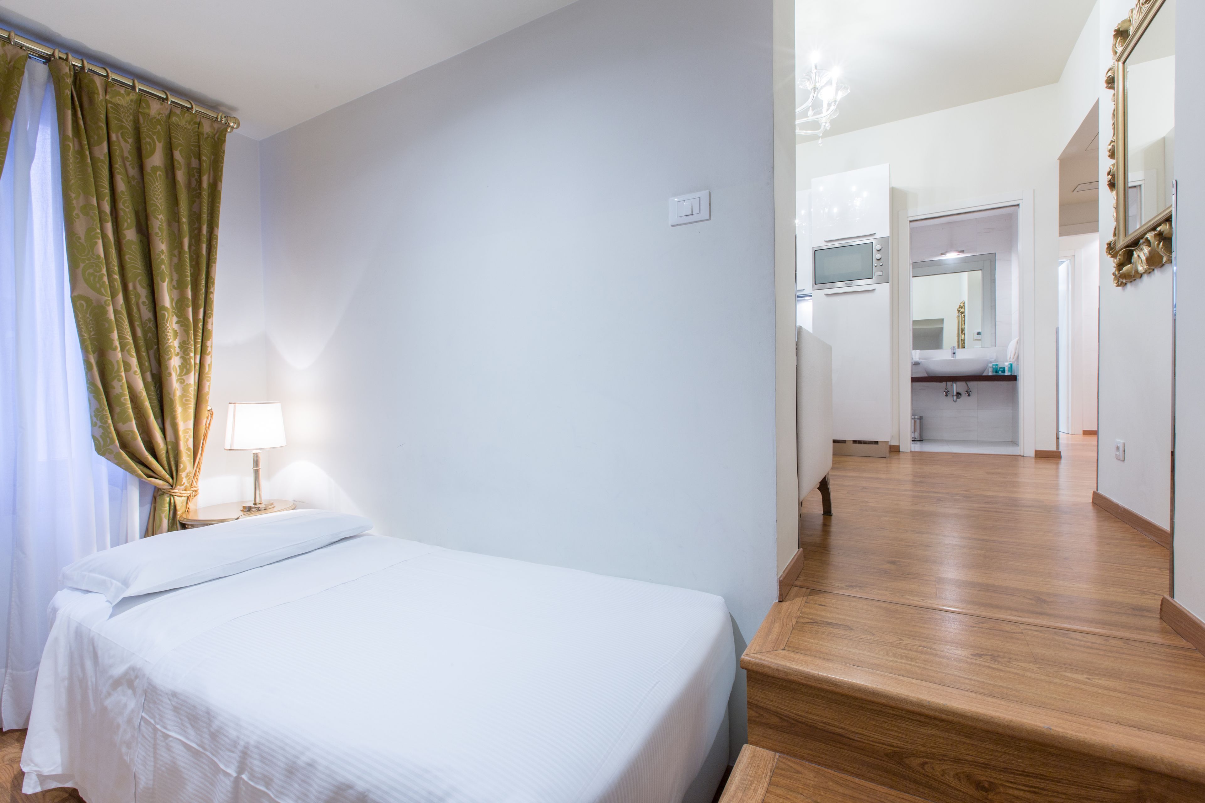 Habitación de hotel con cama, mesita de noche, lámpara, piso de madera, baño con lavamanos y espejo, y una escalera.