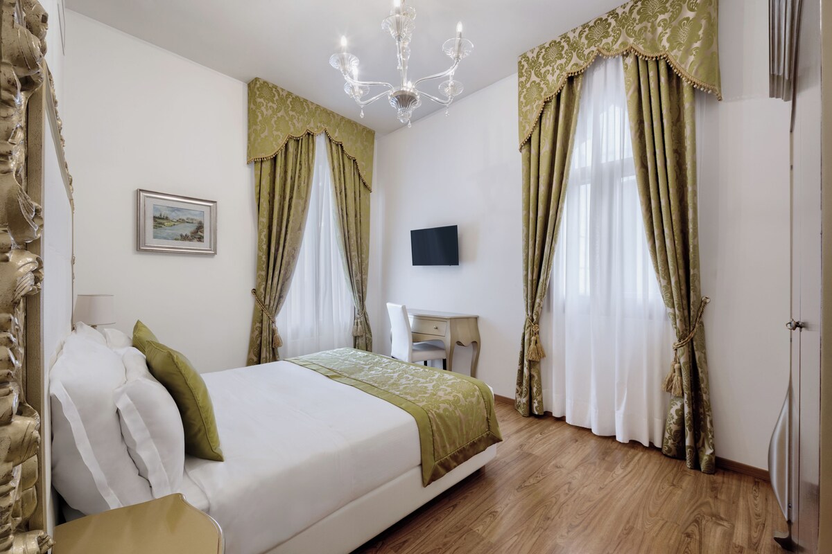 A hotel room with a bed, a desk, a chair, a chandelier, and curtains.