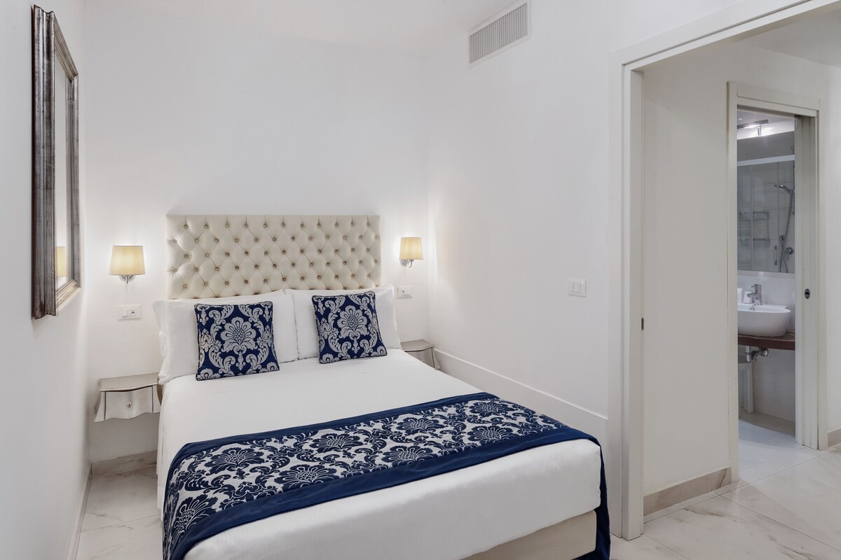 A hotel room with a bed, bedside lamps, a mirror, and a bathroom visible through an open door.