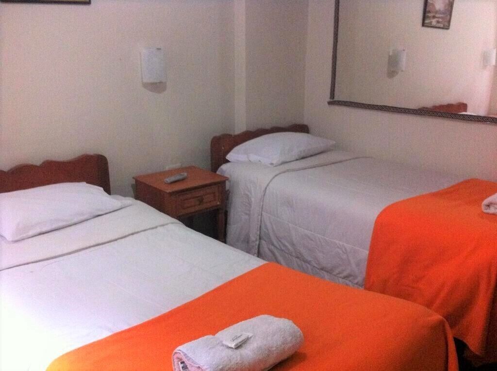Habitación de hotel con dos camas, una con cubrecama naranja y la otra con cubrecama blanca. Entre las camas hay una mesita de noche de madera con un control remoto encima.