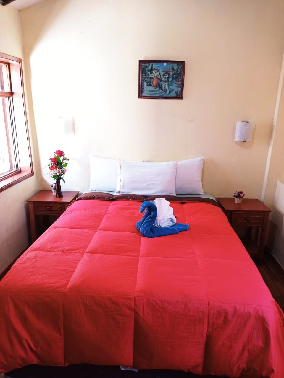 A hotel room with a large bed, a nightstand with a lamp, a painting on the wall, and a window with a view.
