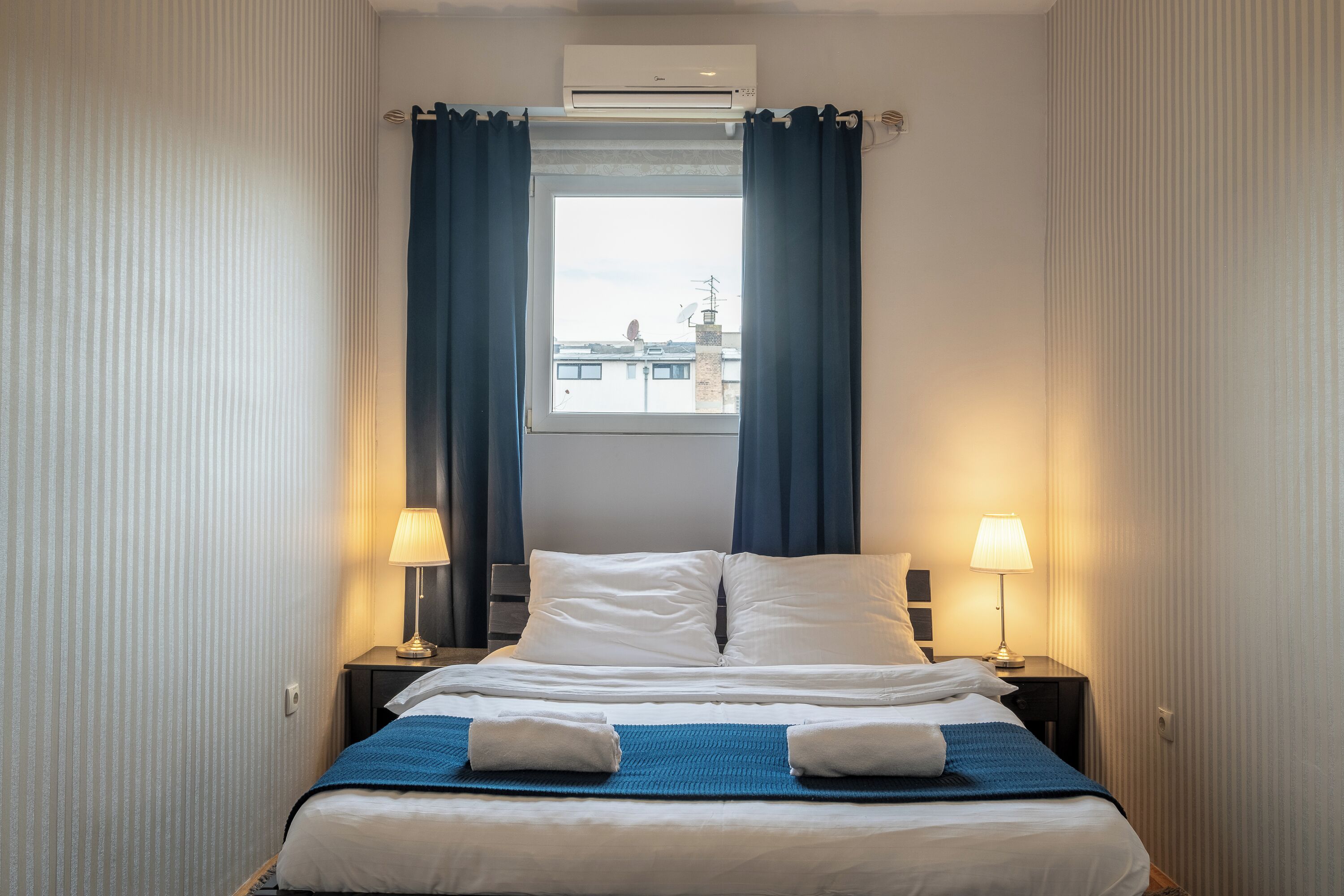 A hotel room with a bed, two bedside lamps, a window with curtains, and an air conditioning unit.