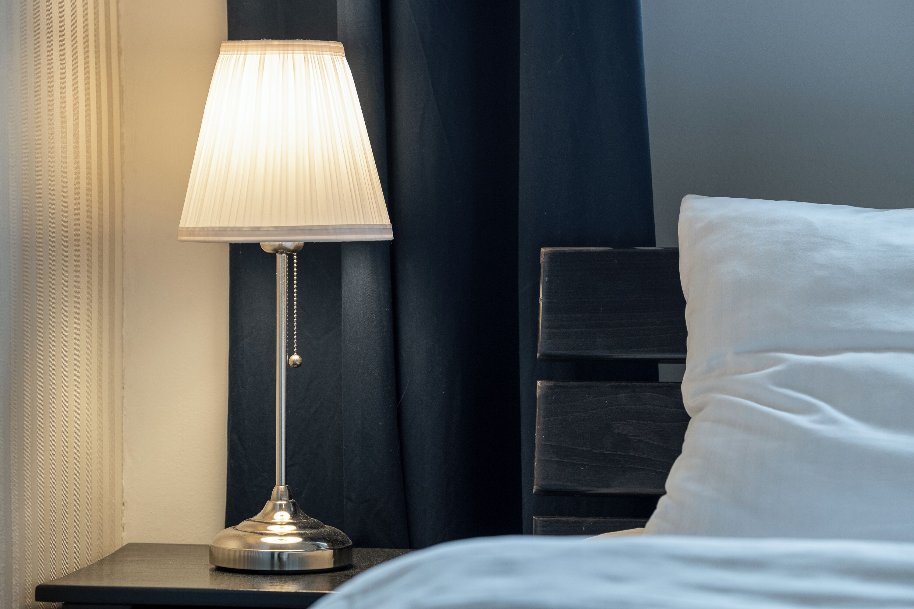 A bed with white linens, a bedside lamp, and a dark-colored headboard.