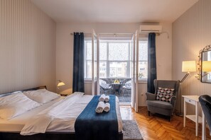 A hotel room with a bed, a desk, a chair, a balcony with a table and chairs, and an air conditioning unit. - Selection Apartments & Rooms (Belgrade)