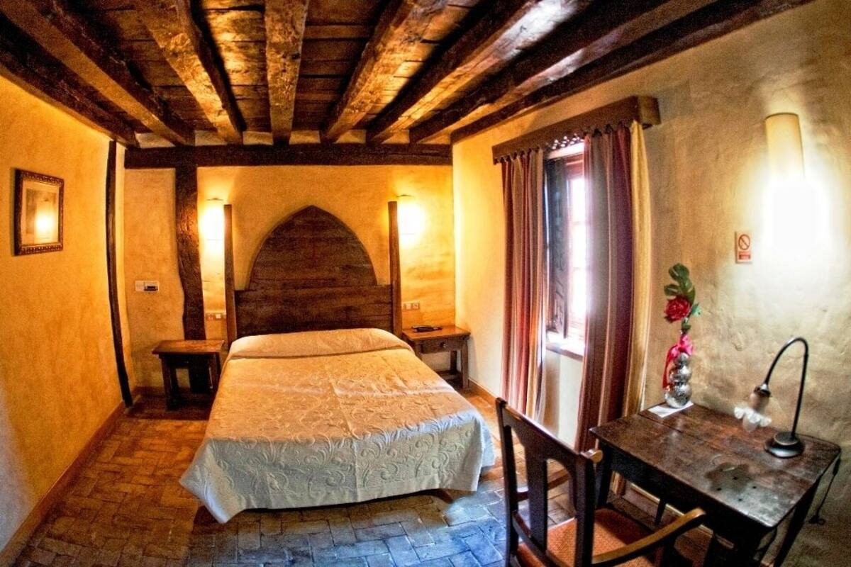 A cozy room with a wooden ceiling, a bed with a white bedspread, a small table with a lamp, and a window with curtains.