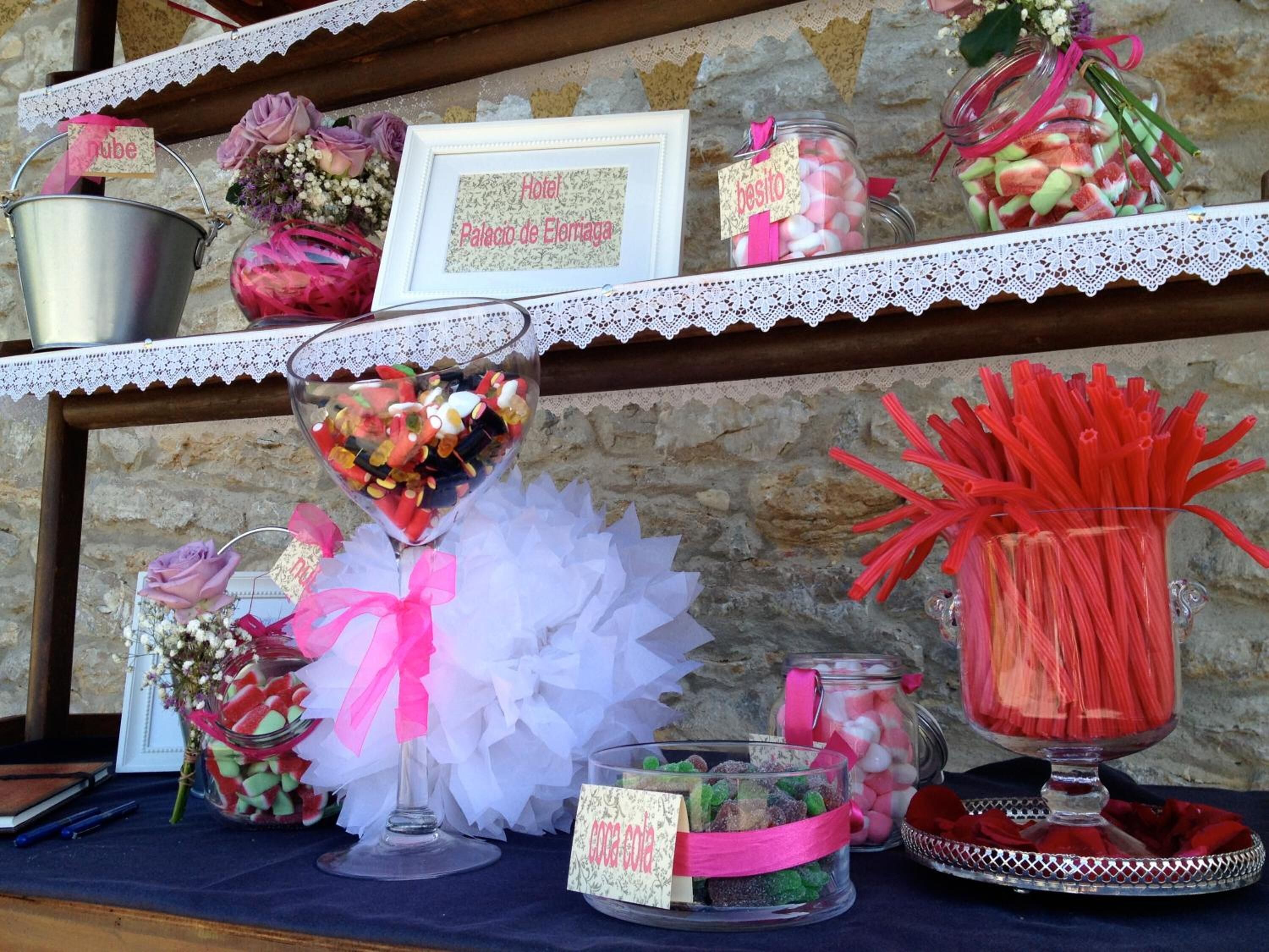 A table with various food items and decorations, including jars of candies and flowers, against a stone wall backdrop.