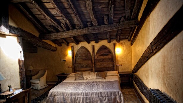 Habitación tradicional con suelo de piedra, vigas de madera, una cama con cubrecama gris, una silla y dos lámparas fijadas a la pared.