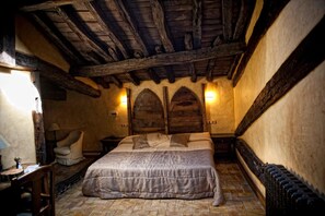 Habitación tradicional con suelo de piedra, vigas de madera, una cama con cubrecama gris, una silla y dos lámparas fijadas a la pared.