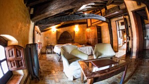 Una habitación rústica con vigas de madera a la vista, paredes de piedra y suelo de piedra. Hay dos camas con sábanas blancas, una mesa de madera y una silla.