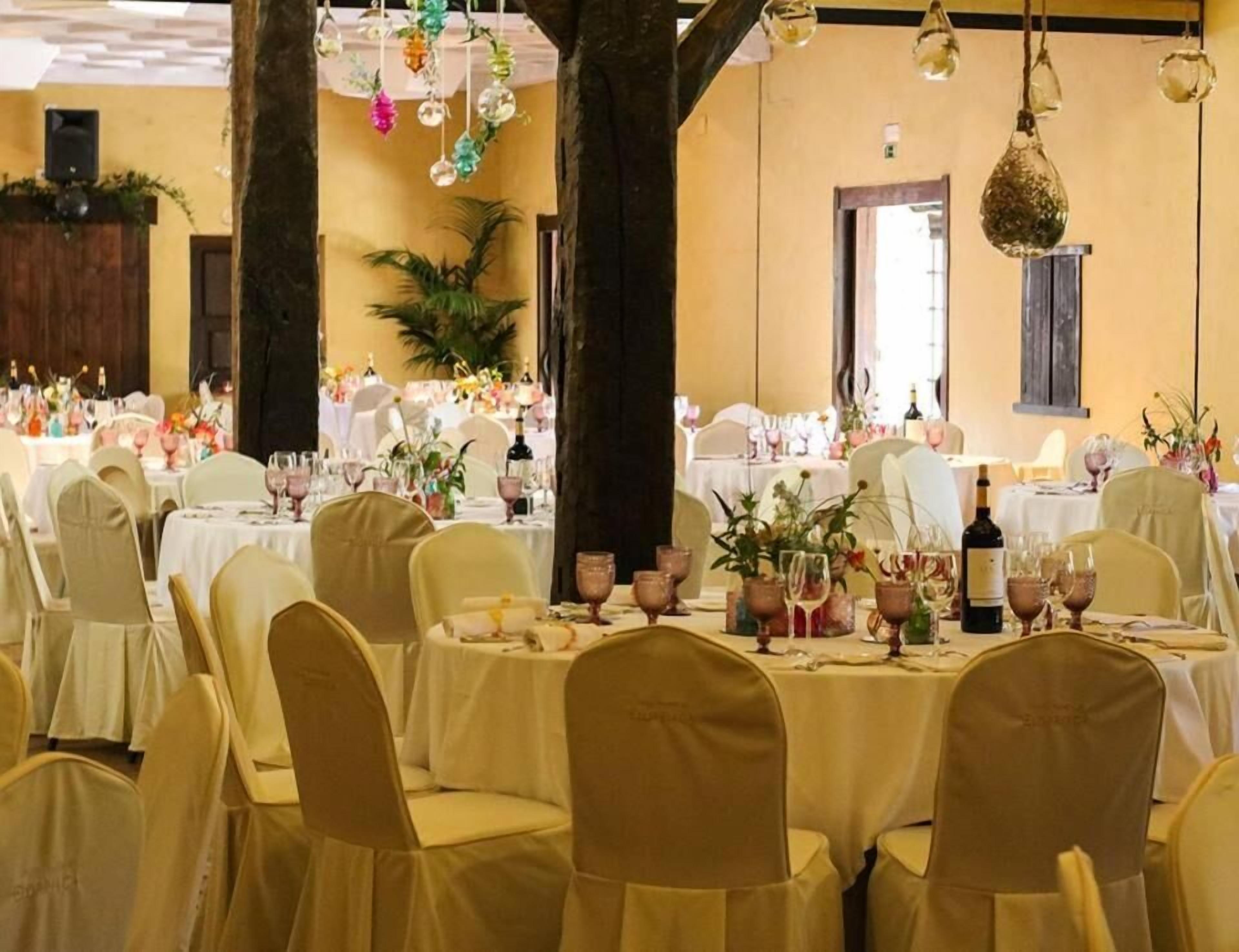 Un salón de banquetes con mesas redondas dispuestas para comer, sillas cubiertas de tela color beige y elementos decorativos colgantes.