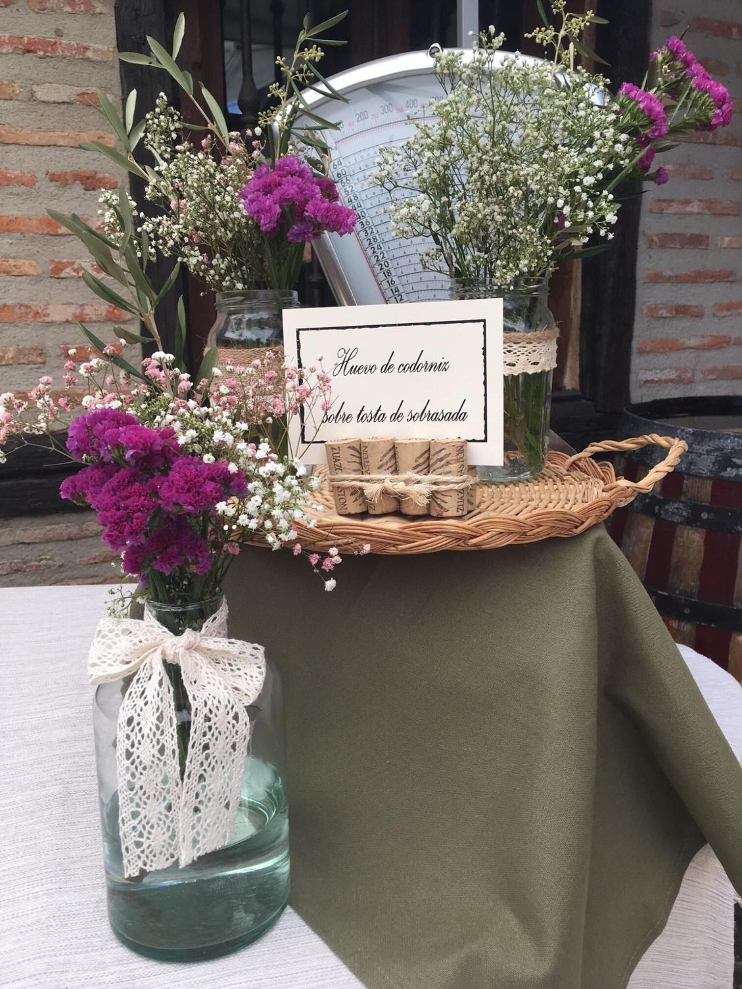 A table with a green tablecloth, decorated with a lace bow, a wicker basket, and two glass jars with lace and flowers.
