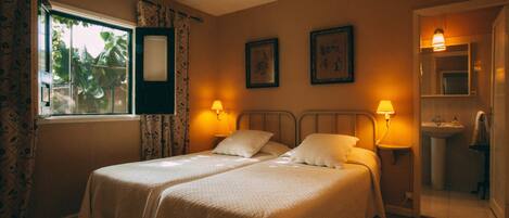 A double bed with two pillows, a bedside table with a lamp, a window with curtains, and a bathroom with a sink and mirror.