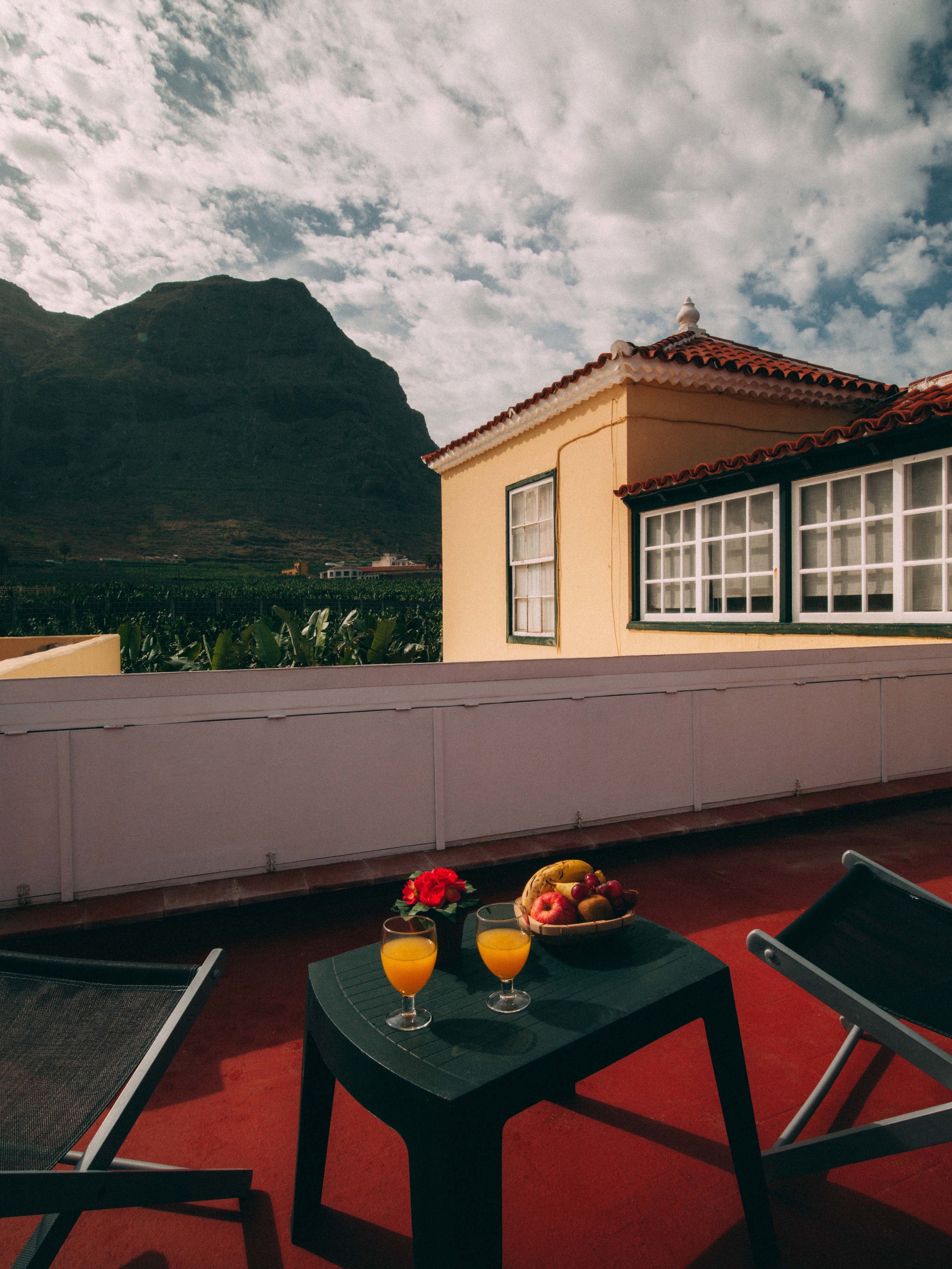 Una terraza con una mesa lista para dos personas, que incluye un frutero y dos vasos de jugo de naranja. Se ve una montaña y un edificio con techo de tejas al fondo.