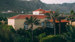 A two-story building with a red-tiled roof, surrounded by lush greenery and palm trees, set against a mountainous backdrop. - Hotel Rural Casamarilla (Garachico)