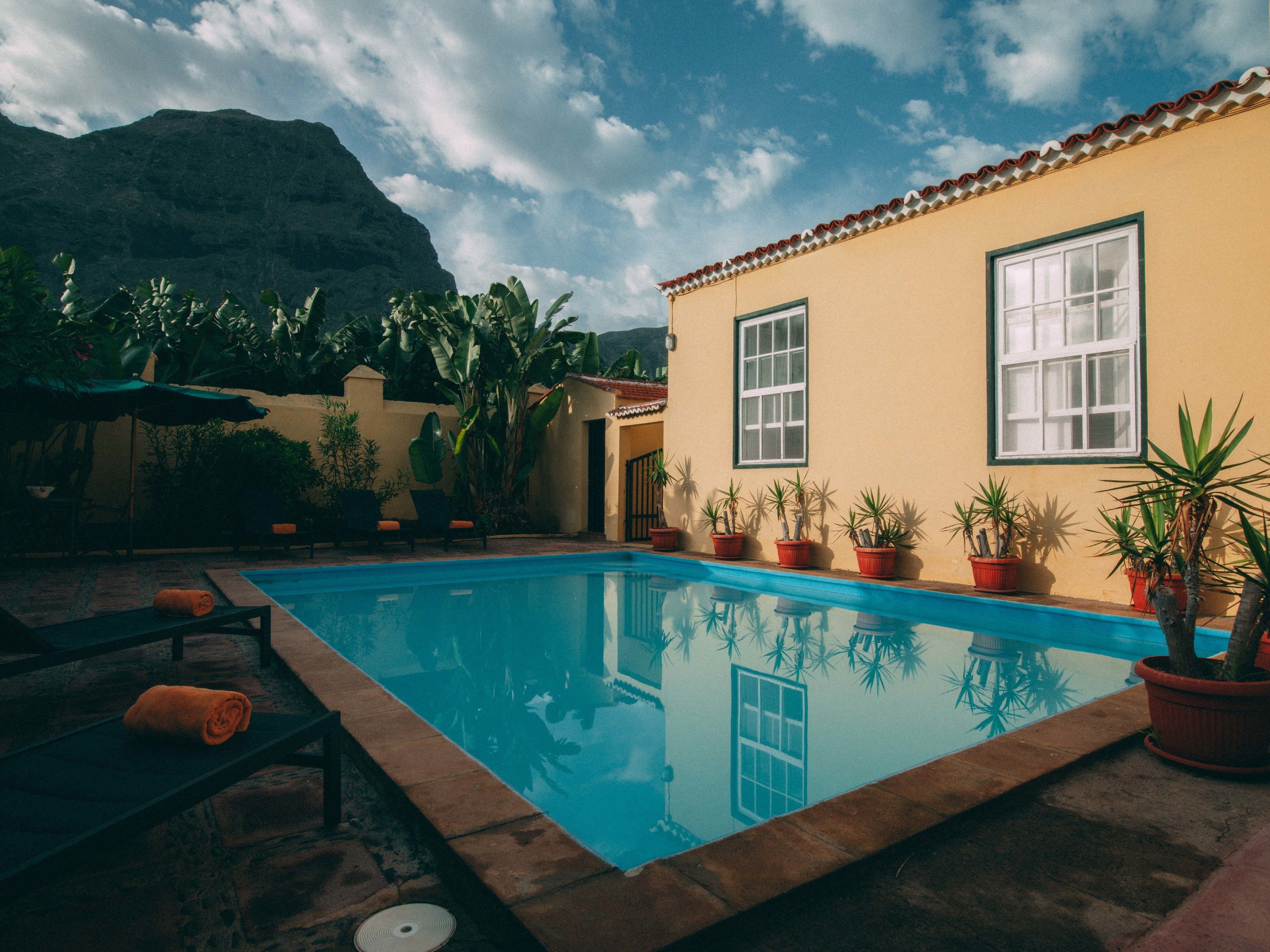 Una piscina con montañas al fondo, plantas en macetas y un edificio con tejado de azulejos.