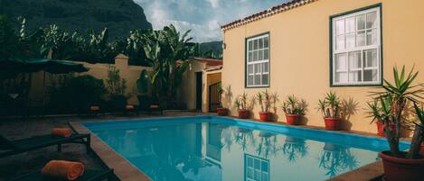 Una piscina con montañas al fondo, plantas en macetas y un edificio con tejado de azulejos.